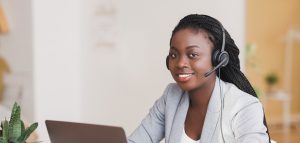 Customer support concept. Young afro female manager wearing headset, using laptop at workplace and looking at camera, free space