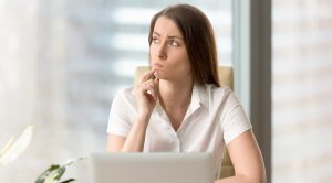 Woman with pensive facial expression looking aside while sitting at workplace. Unsure businesswoman thinking about difficult question. Female office worker doubted because of uncertain situation